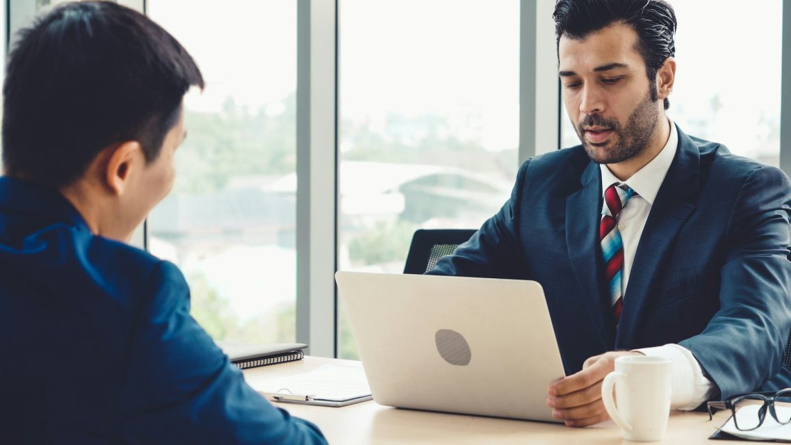 Two men in business attire discussing over a laptop in a bright office setting.