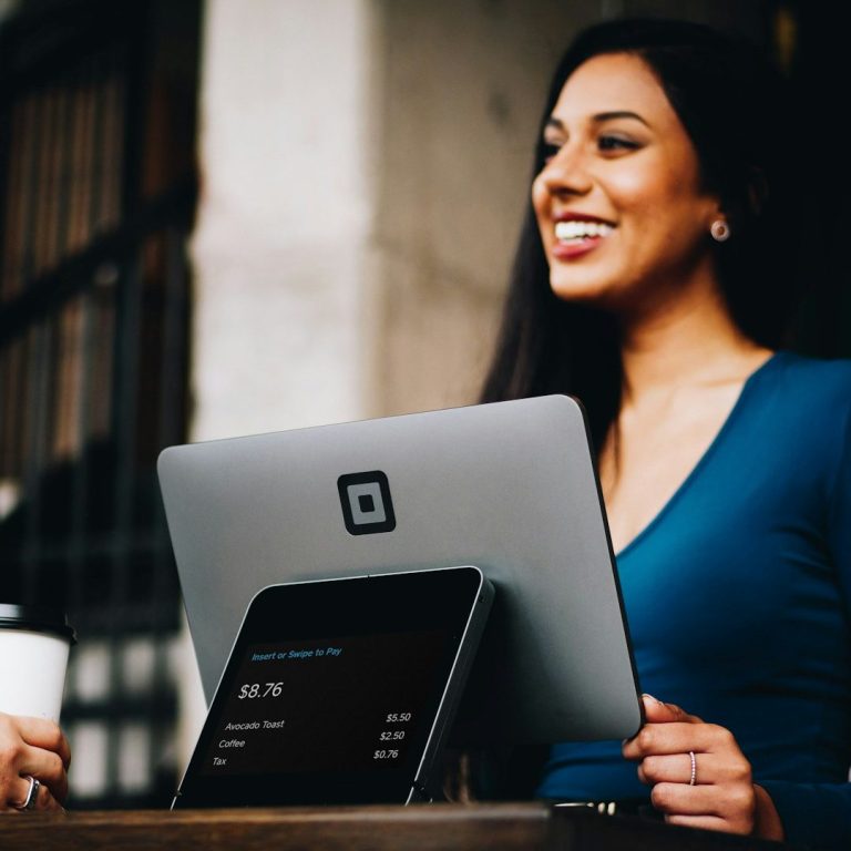Smiling woman in a blue top using a tablet at a café with a coffee cup.