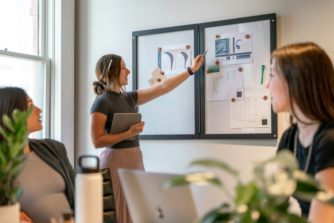 A woman presents ideas on a board to two colleagues in a modern office setting.